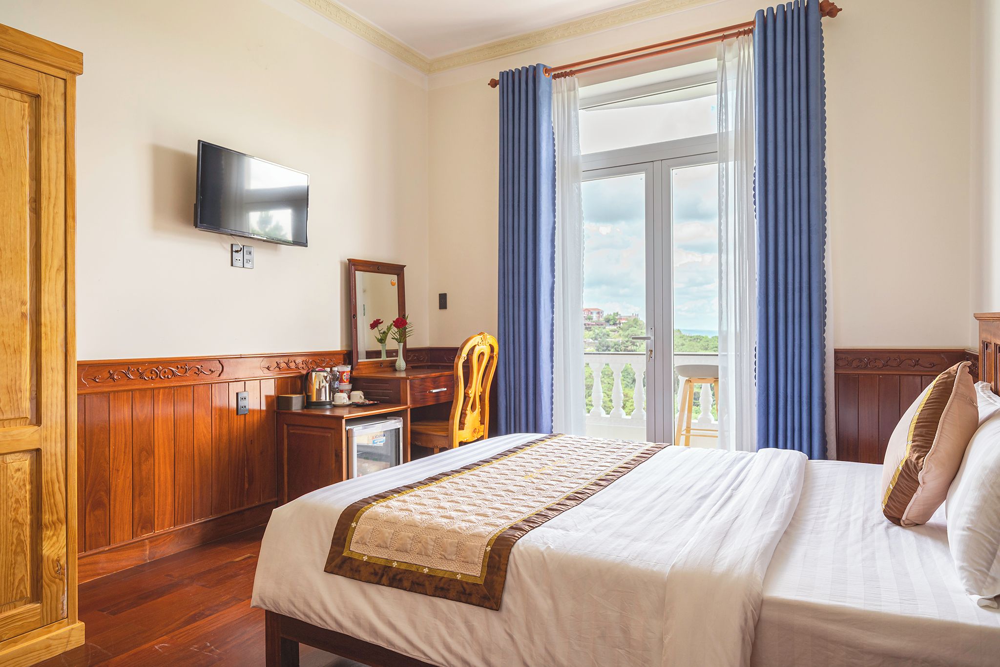 A hotel room with a bed, a desk, a chair, a television, and a balcony with curtains.