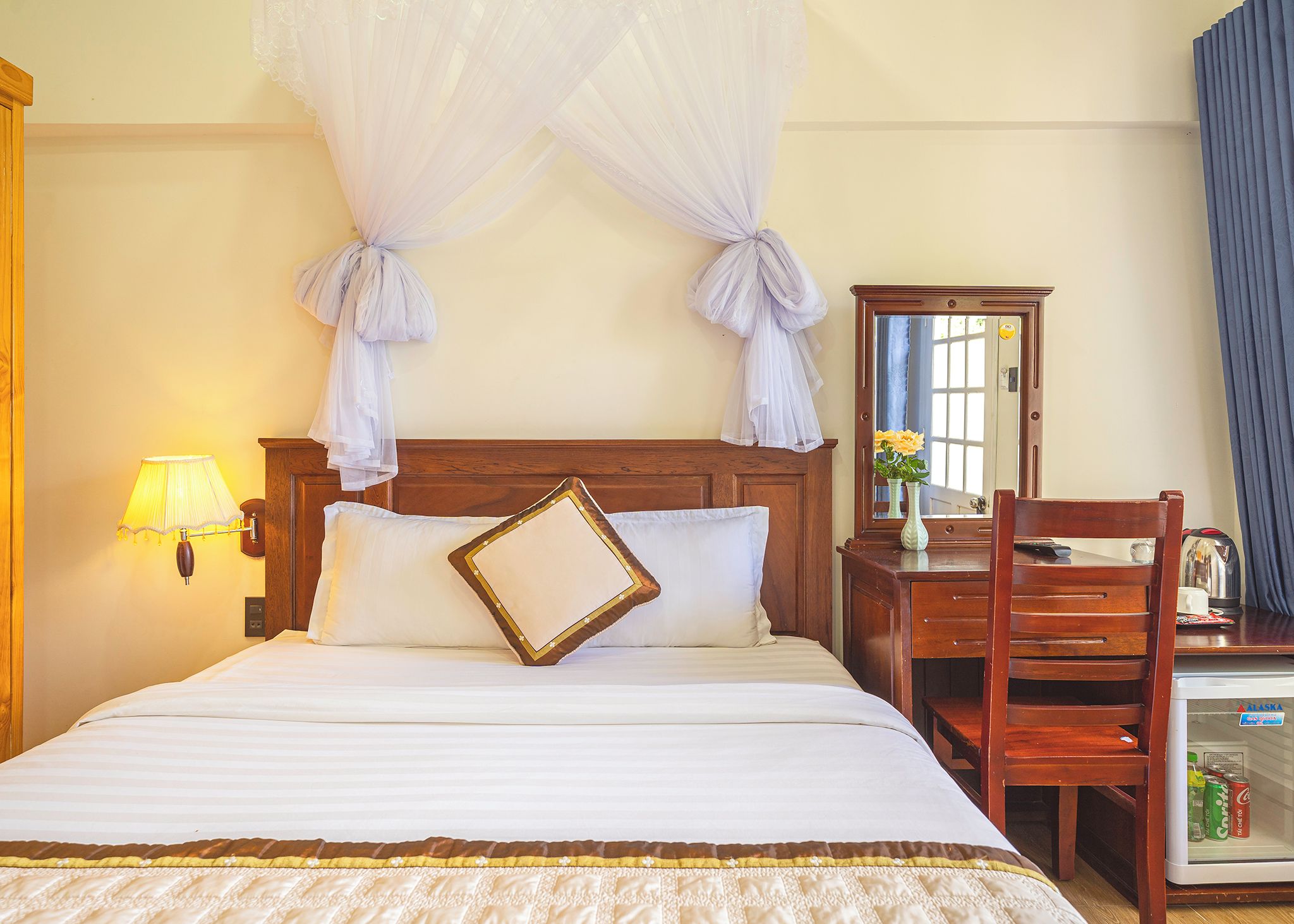 Quarto de hotel com cama, luminária de cabeceira, mesa de cabeceira de madeira, espelho e minibar.