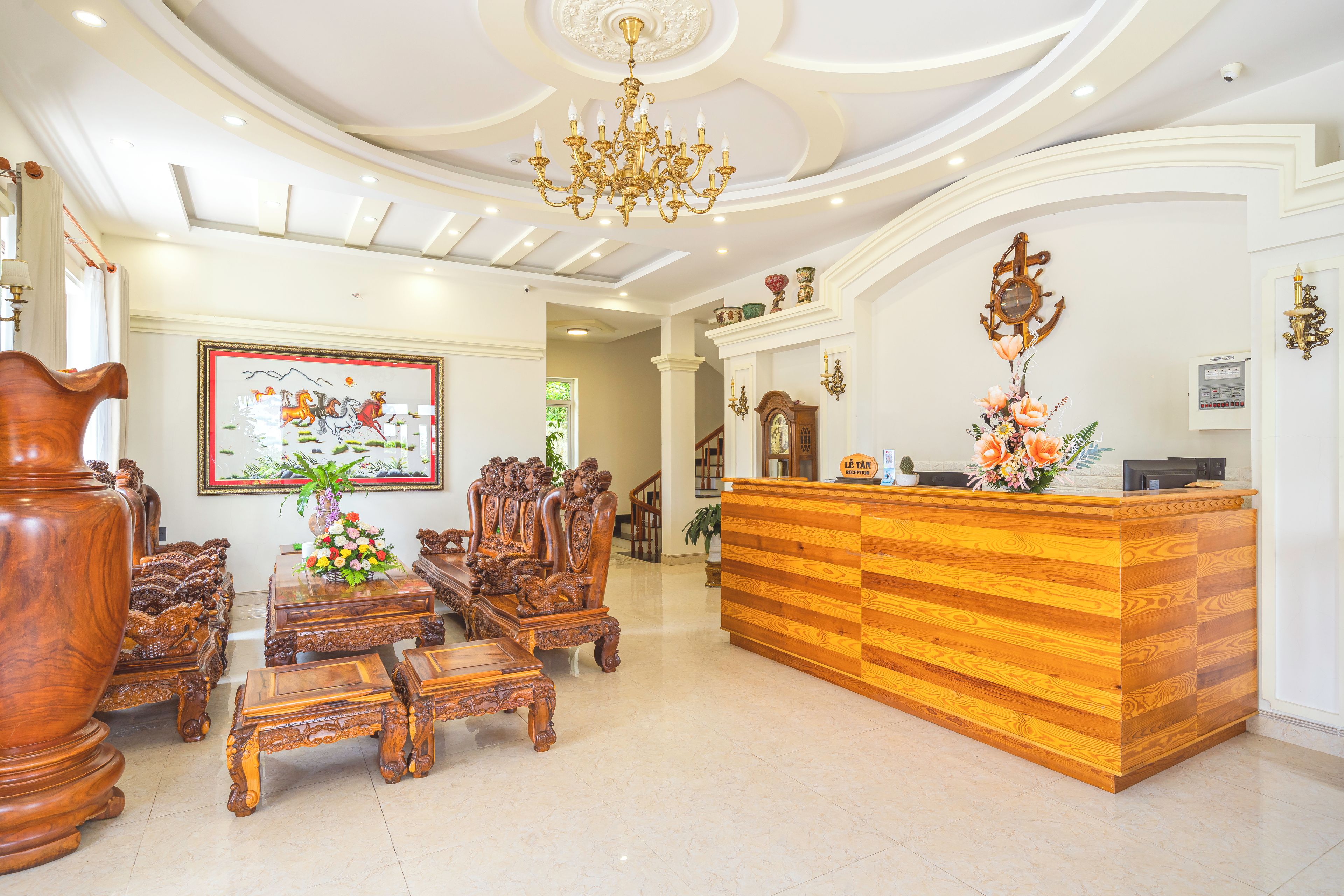 Uma área de recepção com balcão de madeira, cadeiras decoradas, um candelabro e um quadro emoldurado na parede.