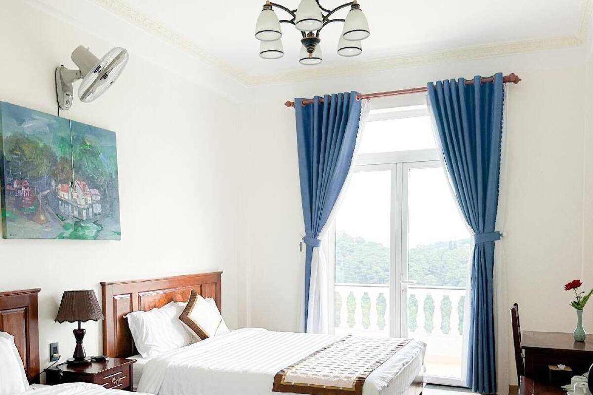 A hotel room with two beds, a ceiling fan, a painting, a chandelier, and a balcony with blue curtains.