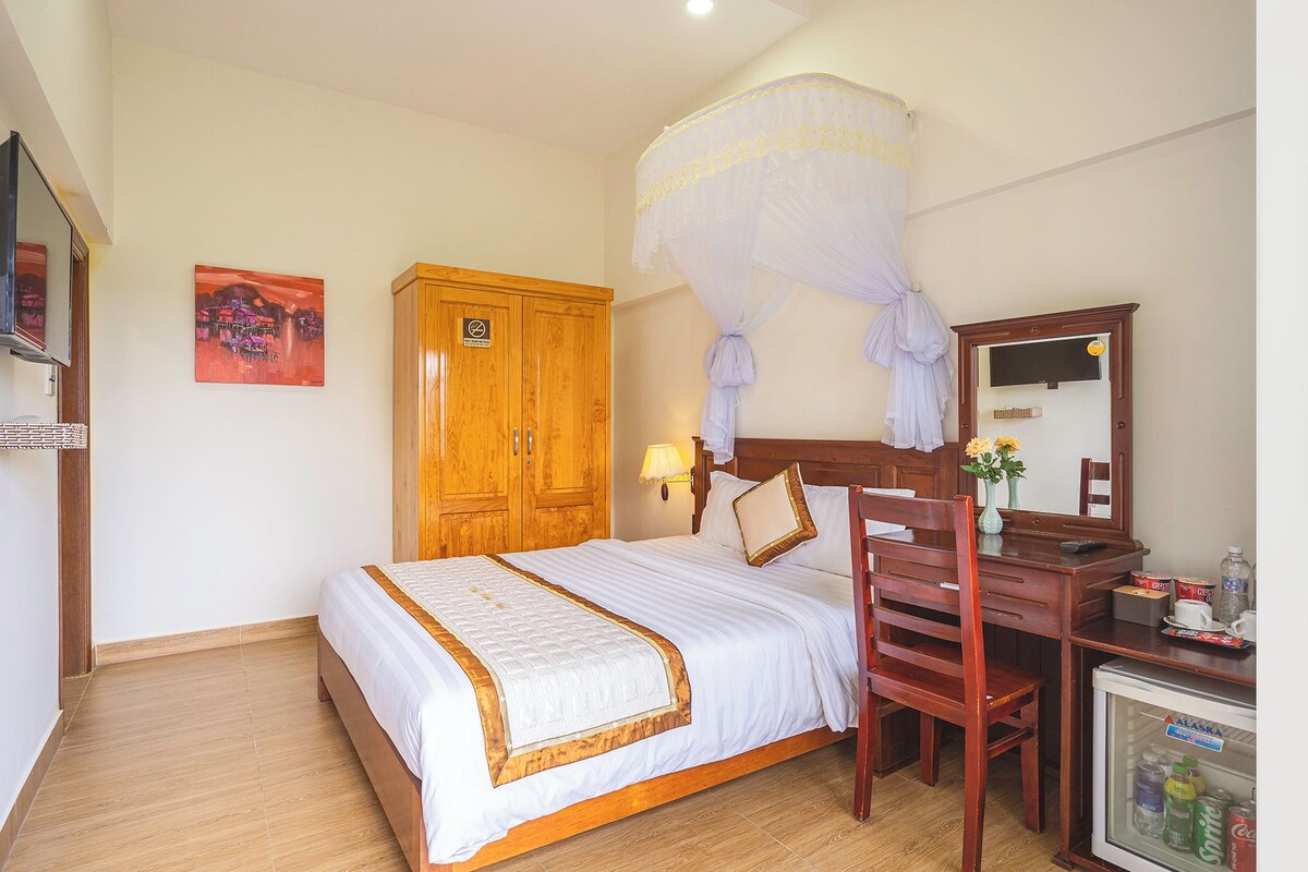 A hotel room with a bed, wooden wardrobe, desk with chair, minibar, and mosquito net.