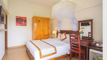 A hotel room with a bed, wooden wardrobe, desk with chair, minibar, and mosquito net.