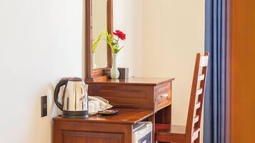 A hotel room with a wooden desk, a television mounted on the wall, a mirror, a vase with a flower, a chair, and a bed with a curtain.