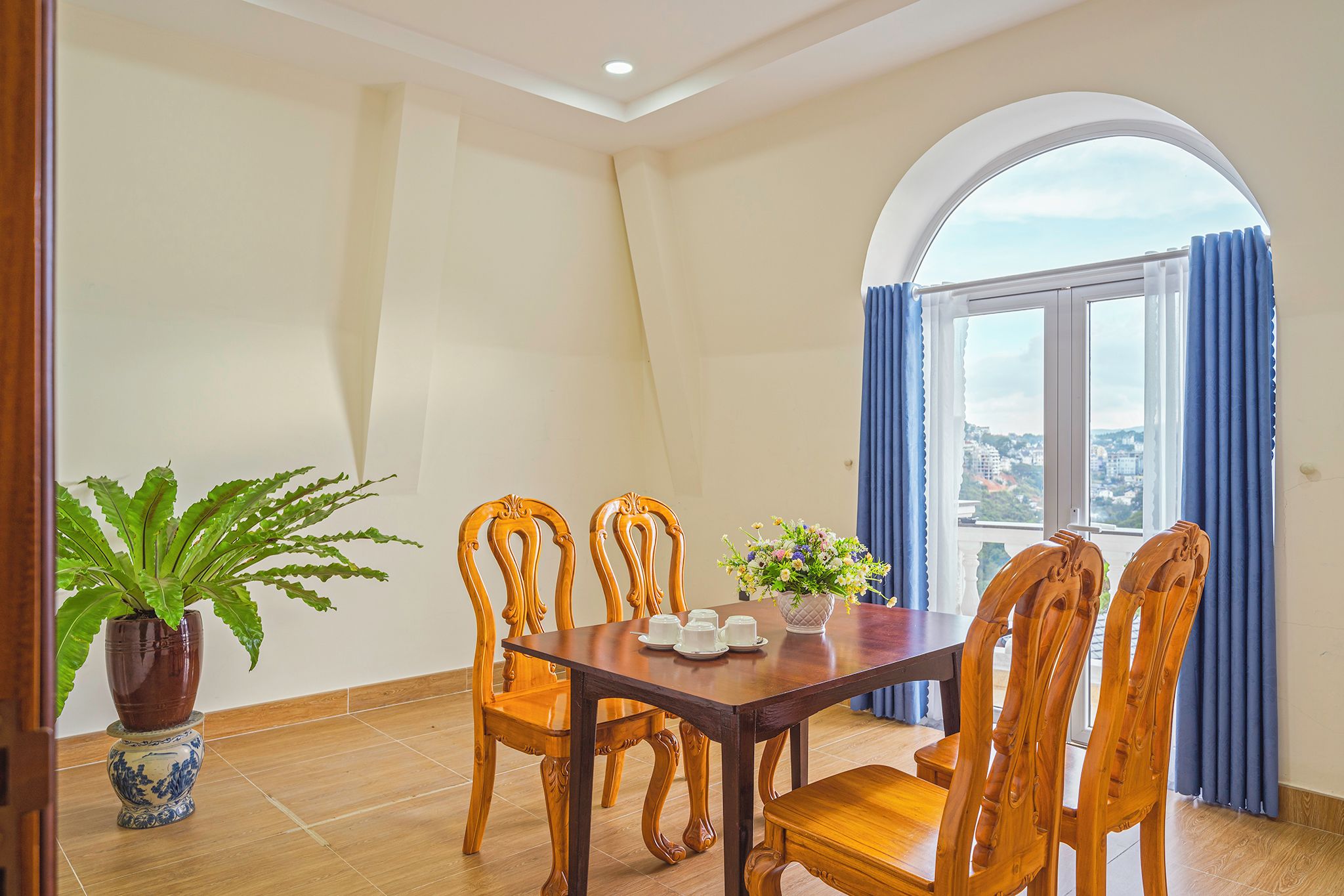 A dining area with a wooden table and chairs, a potted plant, and a view through a window with curtains.