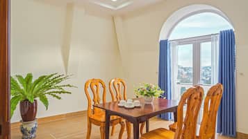 A dining area with a wooden table and chairs, a potted plant, and a view through a window with curtains.
