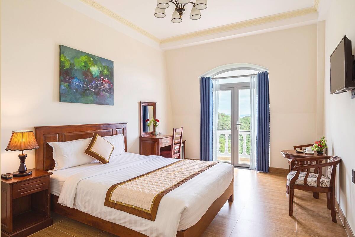 A hotel room with a large bed, a desk with a chair, a TV, and a balcony with curtains.