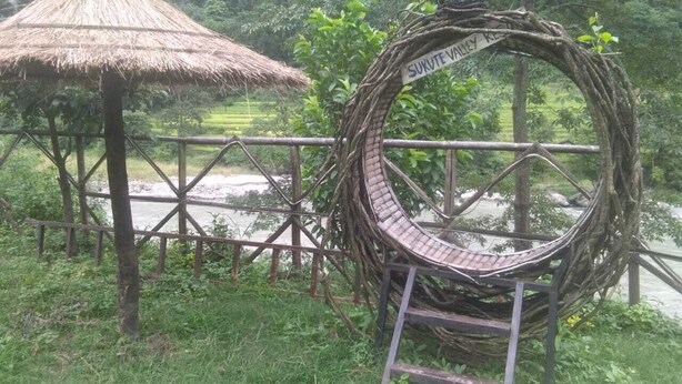 A rustic thatched-roof structure and a circular wooden bridge over a water body, likely a park or natural reserve. - Sukute Valley Resort (Kadambas)