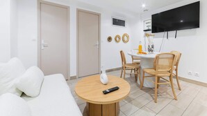 Un salon moderne comprenant un canapé, une table ronde en bois avec une télécommande, un coin repas avec des chaises et un téléviseur fixé au mur.