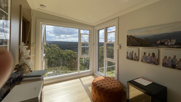 A spacious room with large windows offering a view of a forested area. There is a wooden floor, a tufted ottoman, and a small table with a document on it.