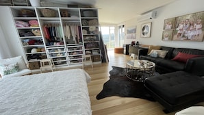 A spacious living room with a large white bed, a black sofa, and a coffee table. There is a white shelving unit filled with various items.