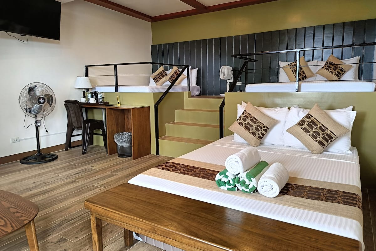 A hotel room with a bed, a desk, a chair, a standing fan, and a staircase.