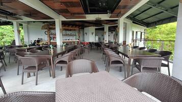 A spacious outdoor dining area with multiple tables and wicker chairs, a bar counter, and a view of greenery.