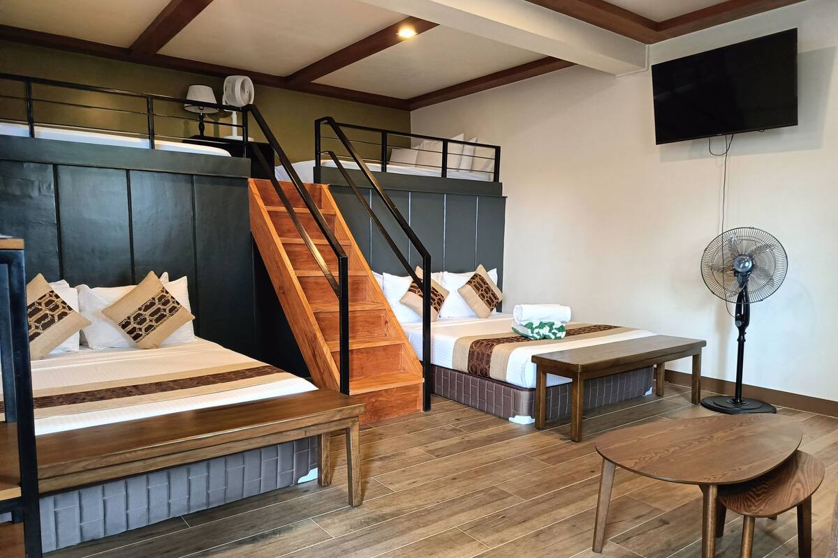 A hotel room with a bunk bed, a desk, a chair, a standing fan, and a TV mounted on the wall.