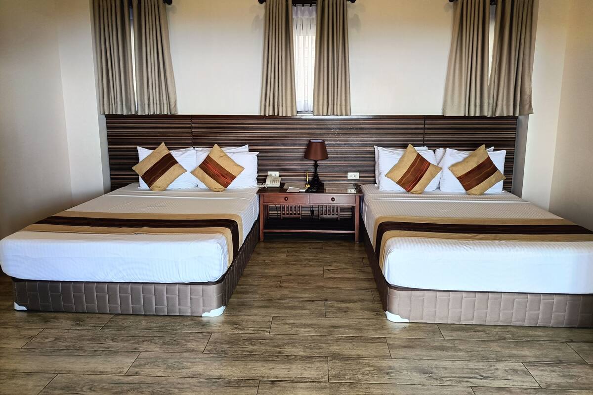 A hotel room with two beds, wooden headboards, beige curtains, and a wooden floor.
