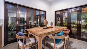 Zona pranzo con tavolo e sedie in legno, una porta vetrata con vista su piante e un ventilatore a soffitto.