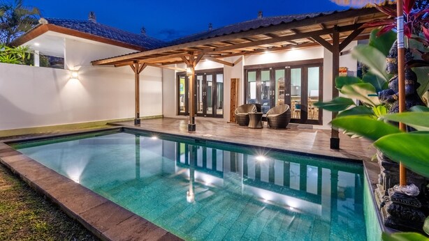 Una piscina con bordo in legno e lettini da relax, circondata da un muro bianco e un'area patio coperta.