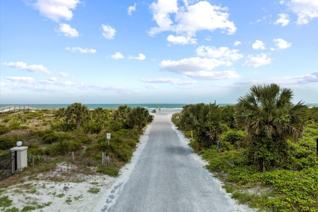 A paved pathway leading to a beach, surrounded by greenery and palm trees.