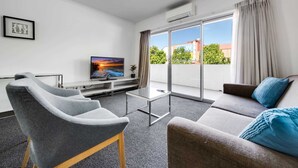 A modern living room with a flat-screen TV, grey armchairs, a sofa with a blue throw pillow, and a sliding glass door leading to a balcony.