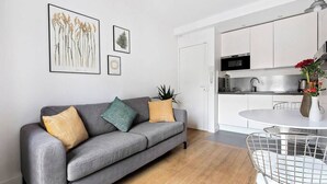 A modern living room with a grey sofa, a round dining table, and a kitchen area visible through an open door.