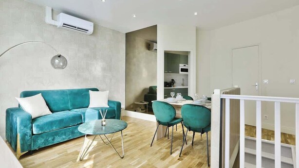 Una sala moderna con un sofá azul aguamarina, una mesa redonda de vidrio y sillas azul aguamarina. Se ve un área de cocina a través de un vano abierto.