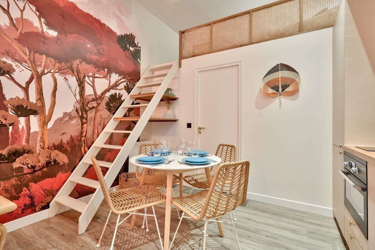 A dining area with a round table set for four, surrounded by wicker chairs, a staircase, and a wall mural.