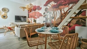 A modern living room with a dining area, featuring wicker furniture, a large mural, and a staircase.