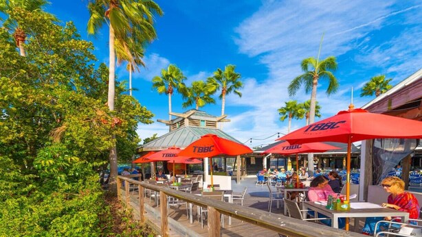 A tropical outdoor dining area with red umbrellas labeled "TBBL" and a wooden deck.