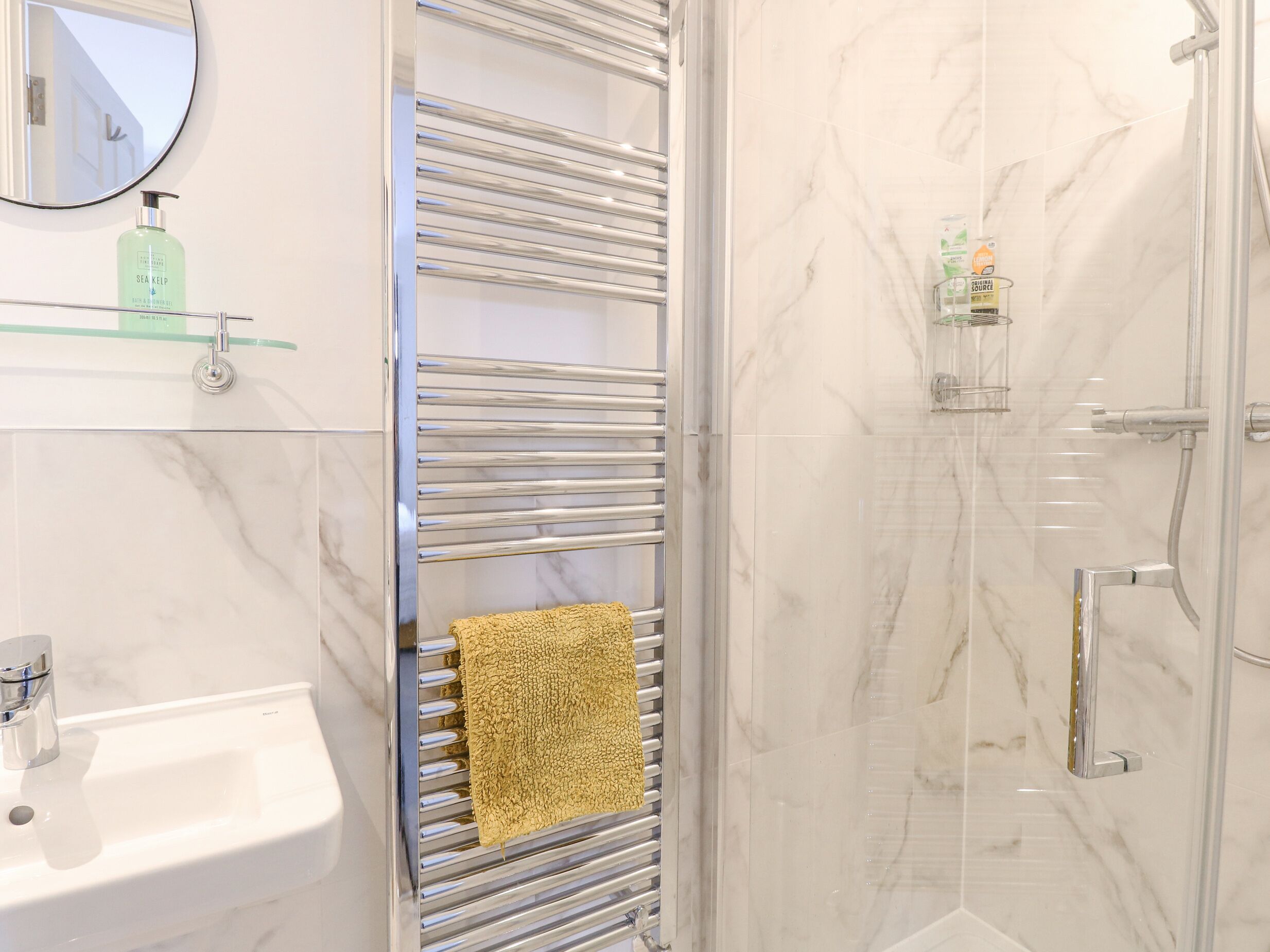 A bathroom with a marble sink, a towel rack with a yellow towel, a glass shower enclosure, and a mirror with a soap dispenser.