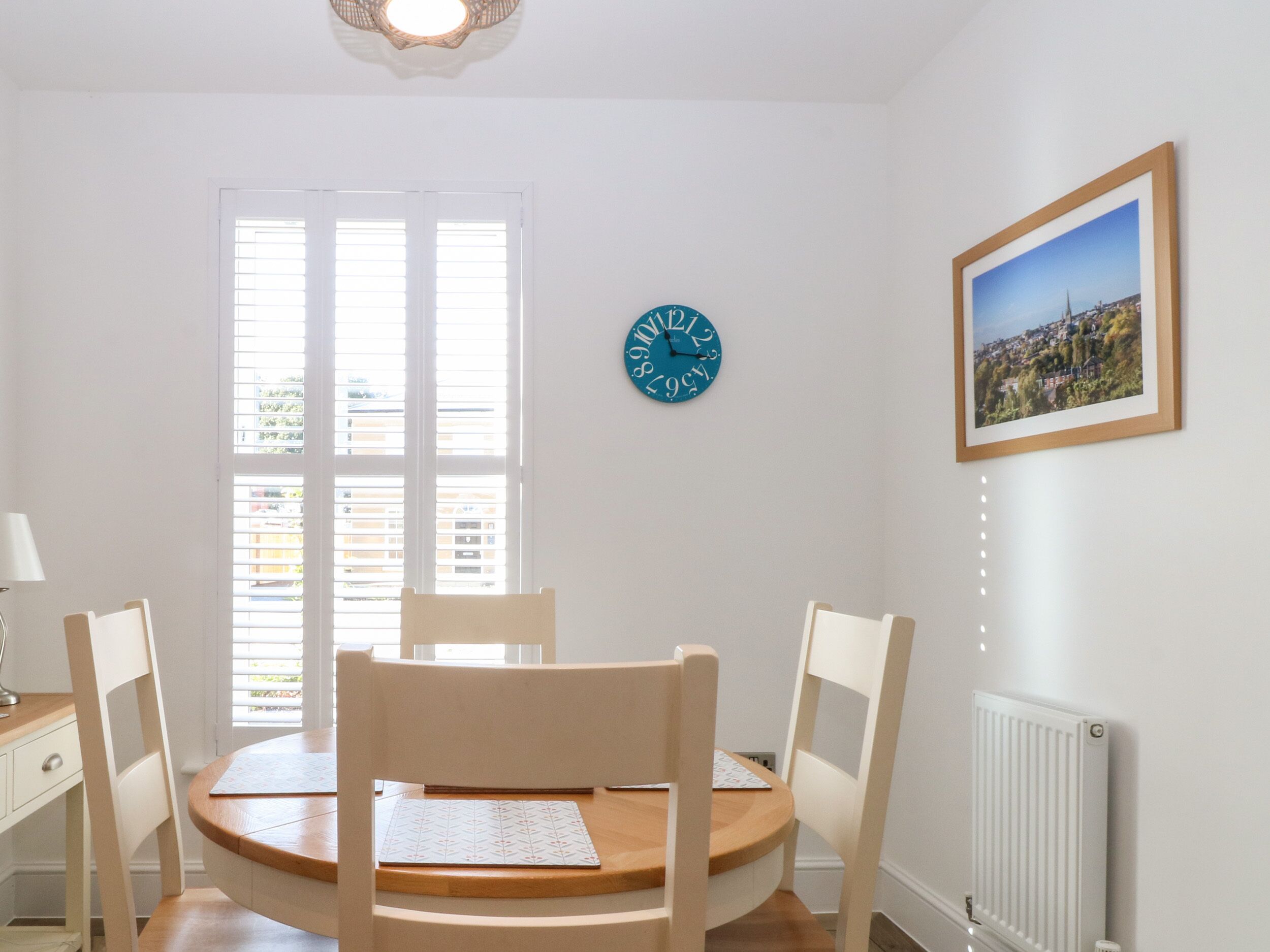 A dining area with a round table, four chairs, a wall clock, a framed picture, and a window with blinds.