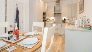 A modern kitchen with white cabinets, a wooden dining table set for four, and a small island with a coffee maker. - The Hollies (Norwich)