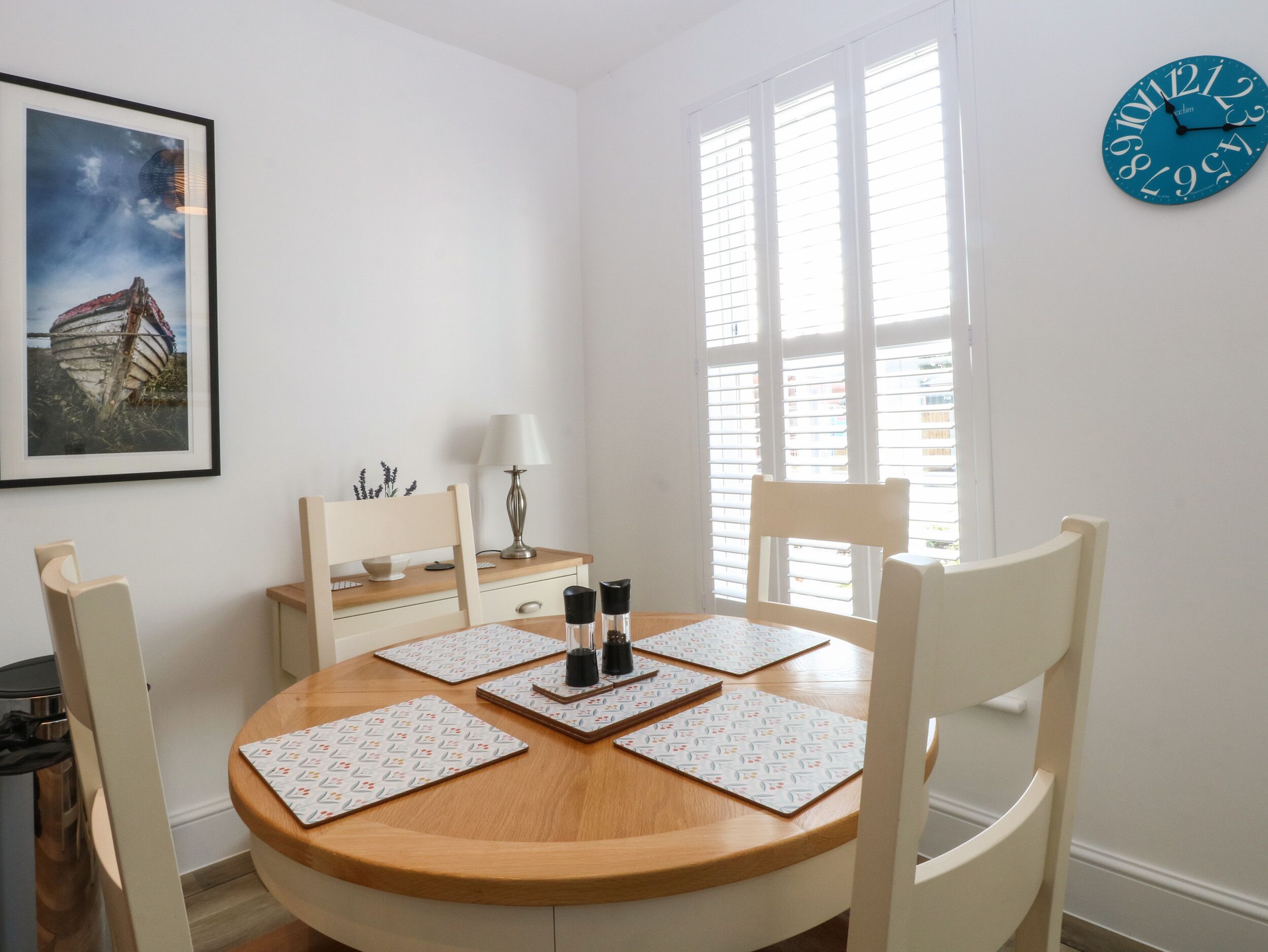 A dining area with a round wooden table, white chairs, a framed picture on the wall, a lamp, and a window with blinds.