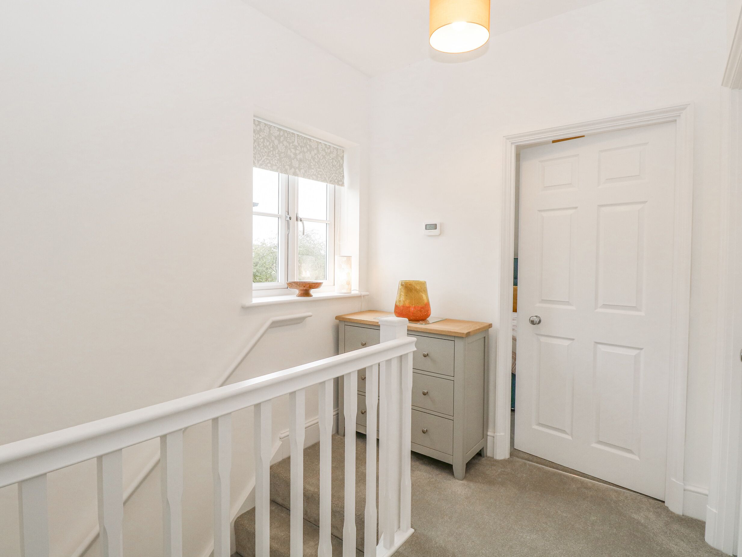 A narrow hallway with a white door, a window with a decorative valance, a small table with a glass container, and a white railing.