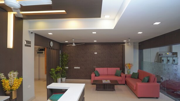 Un área de recepción moderna con un mostrador blanco, un sofá rojo con cojines verdes, una planta en maceta y un ventilador de techo.