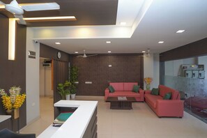 A modern reception area with a white reception desk, a red sofa with green cushions, a potted plant, and a ceiling fan. - HOTEL ZAIN GRANDE (Chennai)
