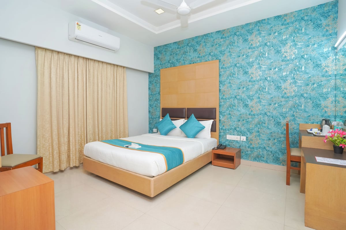 A hotel room with a bed, a desk, a chair, a nightstand, an air conditioning unit, and a window with curtains.