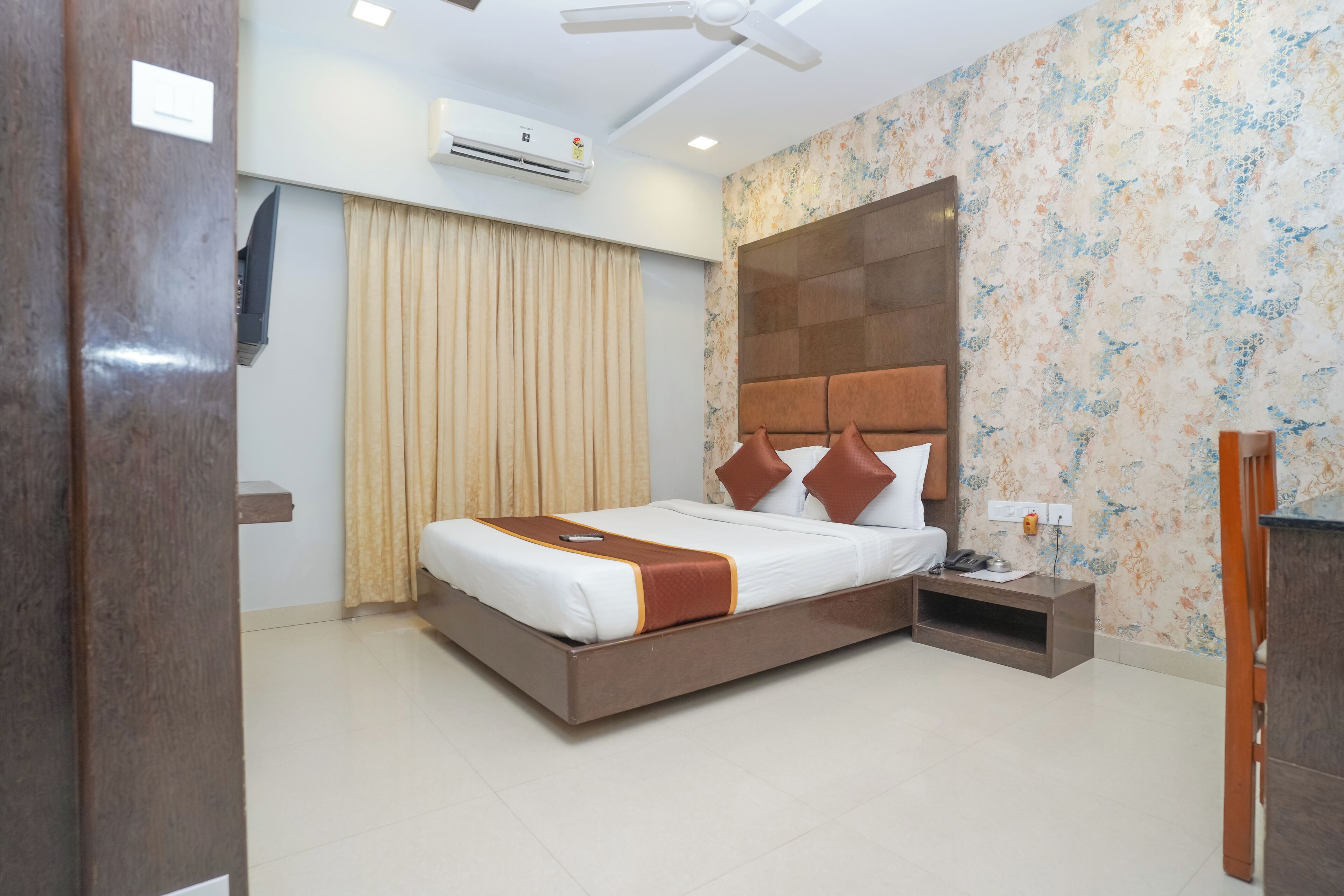 A hotel room with a large bed, a nightstand, a ceiling fan, an air conditioning unit, and a decorative wall panel.