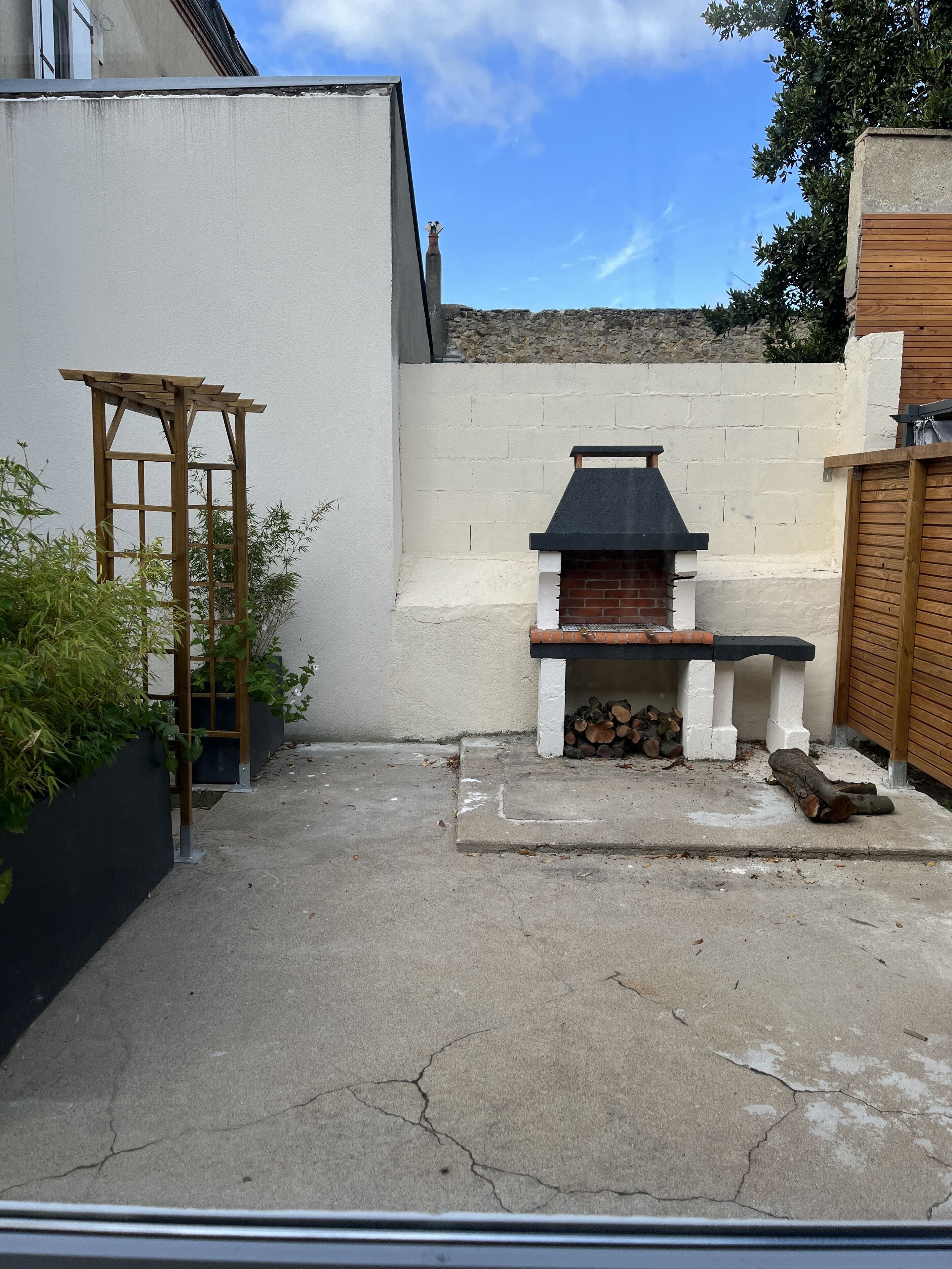 A backyard with a brick barbecue, a wooden pergola, and a concrete floor.