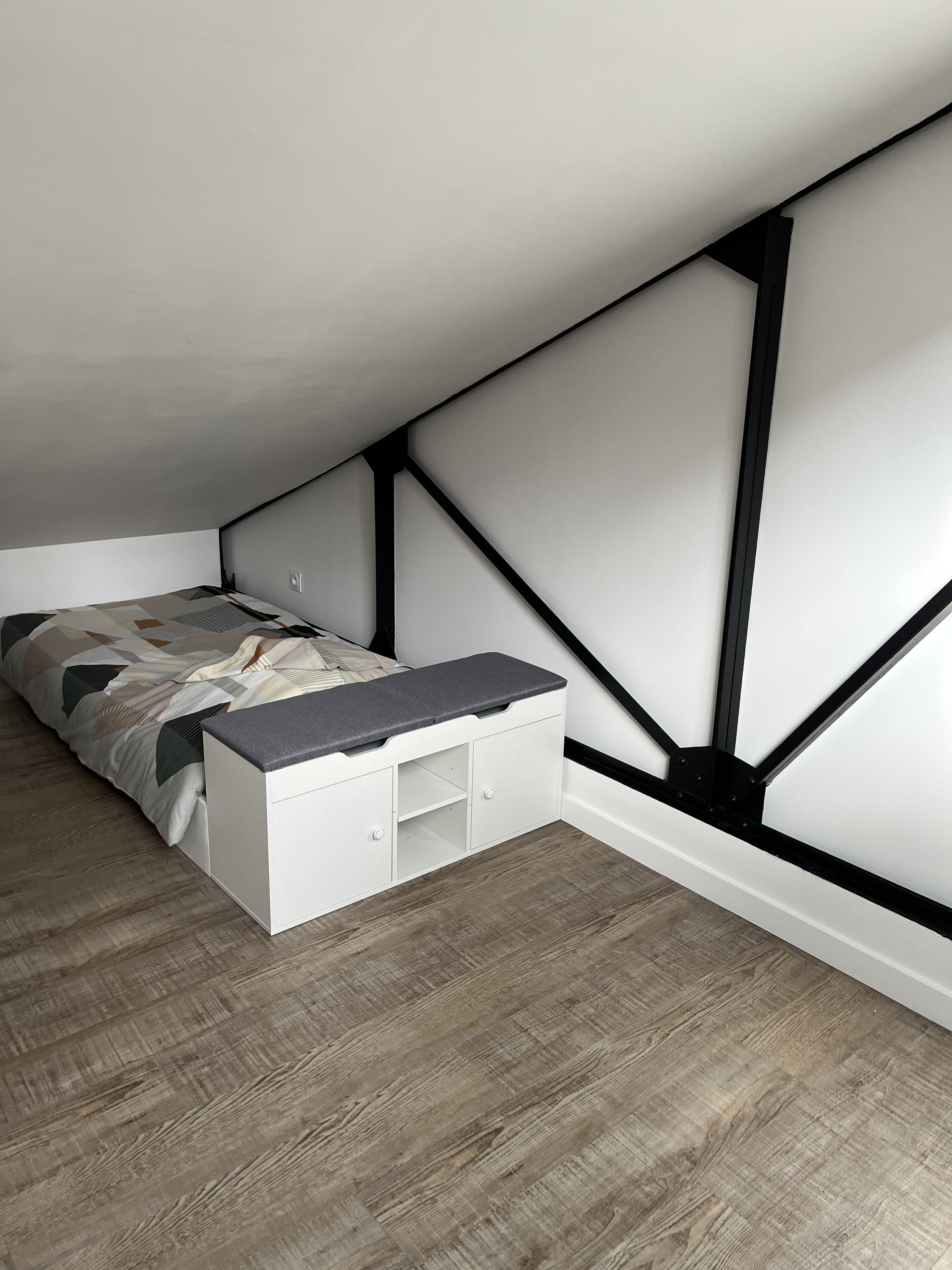 A bedroom with a sloped ceiling, a bed with a geometric-patterned duvet, a white storage unit with drawers, and a wooden floor.