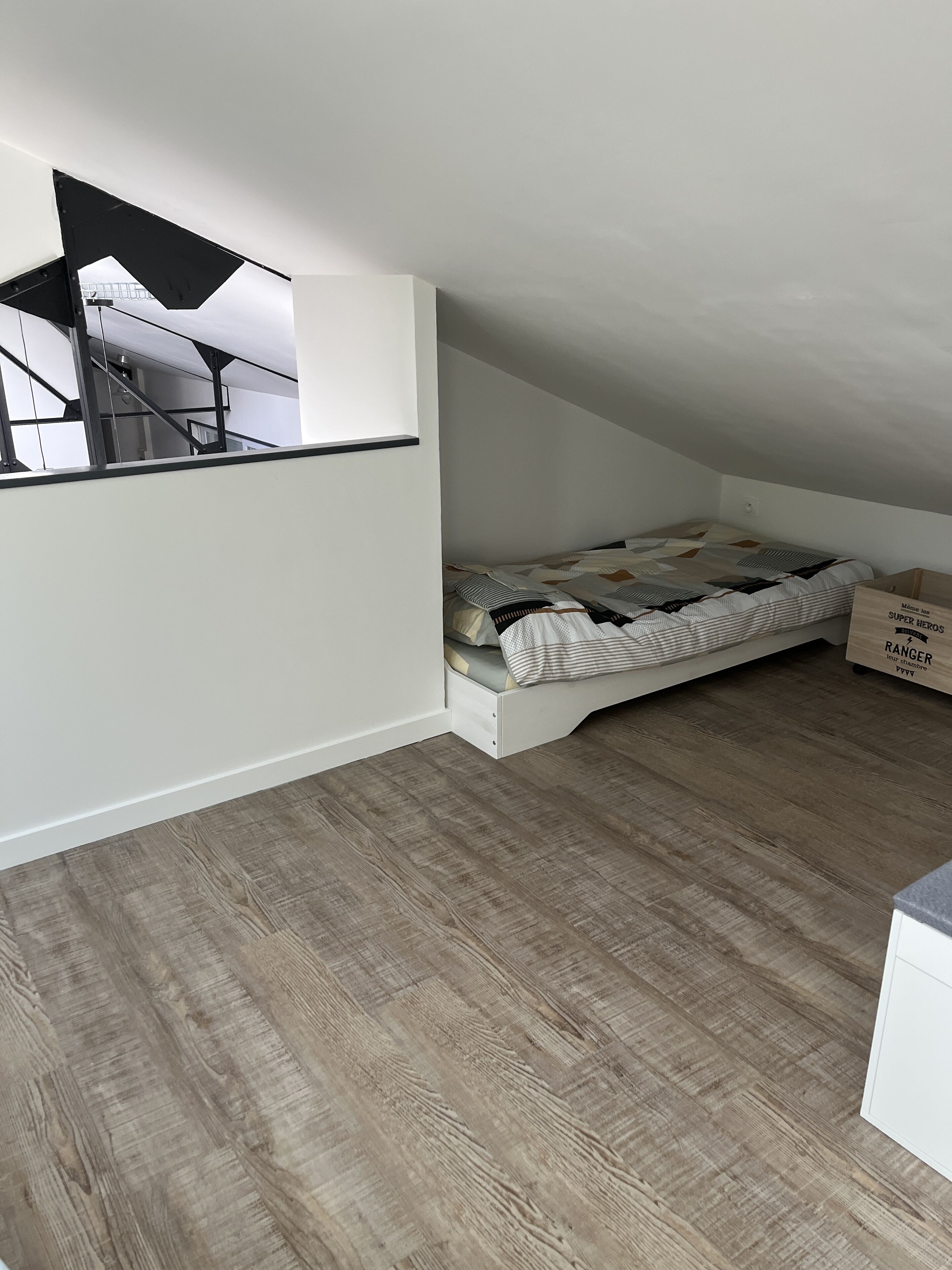 A small, sloped ceiling room with a bed, a white wall, and a wooden floor.