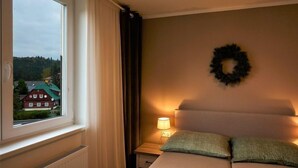 A bedroom with a bed, a nightstand with a lamp, a window with curtains, and a wreath on the wall. - Harrachov 377 by Interhome (Harrachov)