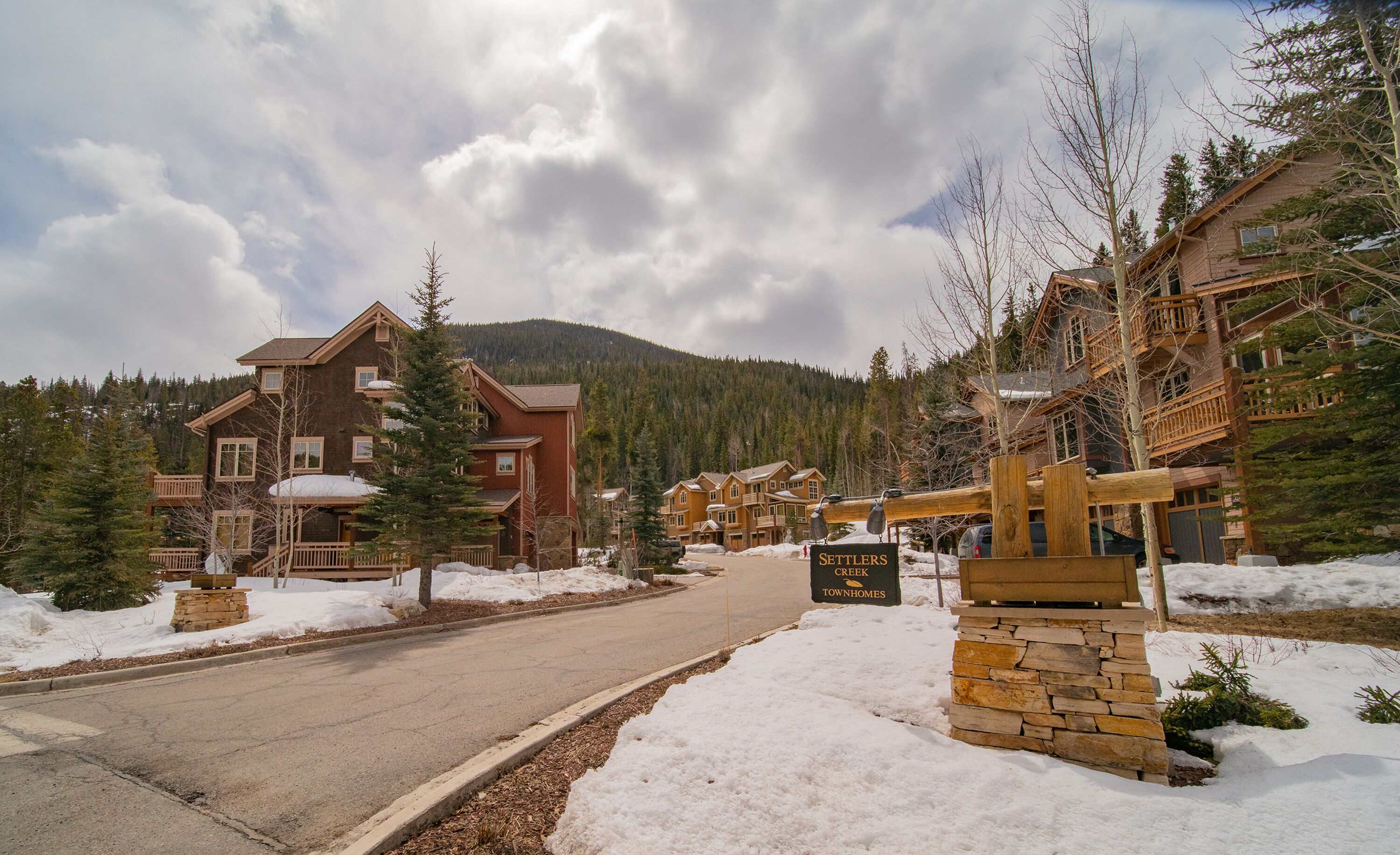 A snow-covered street leading to a lodge with multiple buildings, surrounded by trees and mountains.