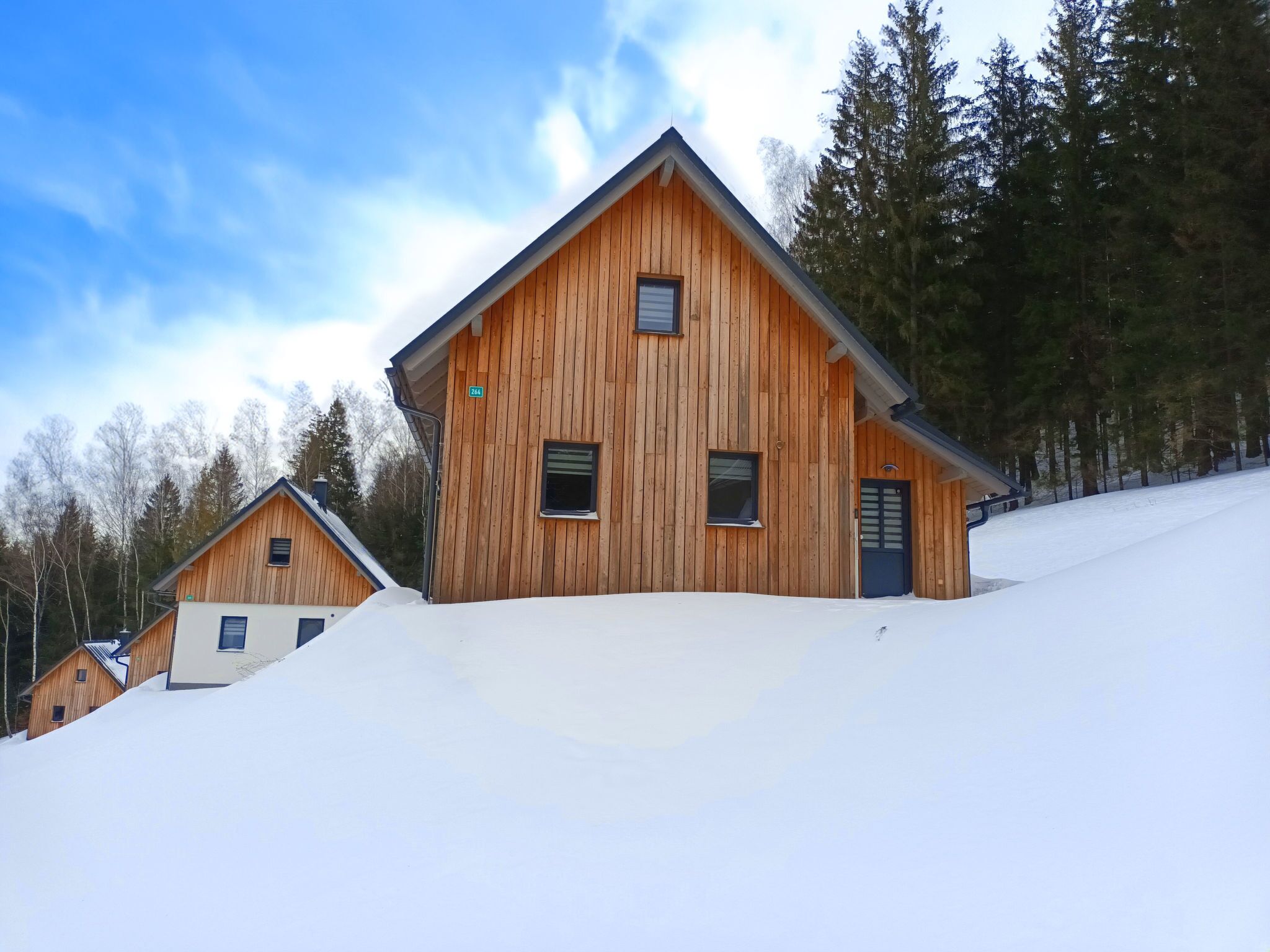 A wooden cabin with a steep roof, surrounded by snow-covered ground and trees.