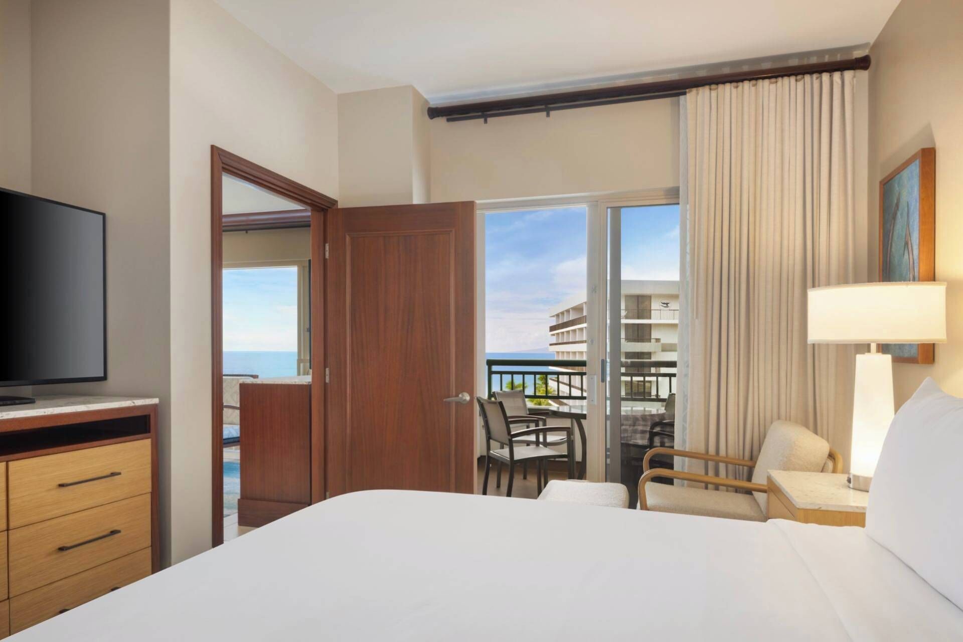 Quarto de hotel com cama, televisão, secretária com gavetas, cadeira e varanda com vista para edifícios e o mar.