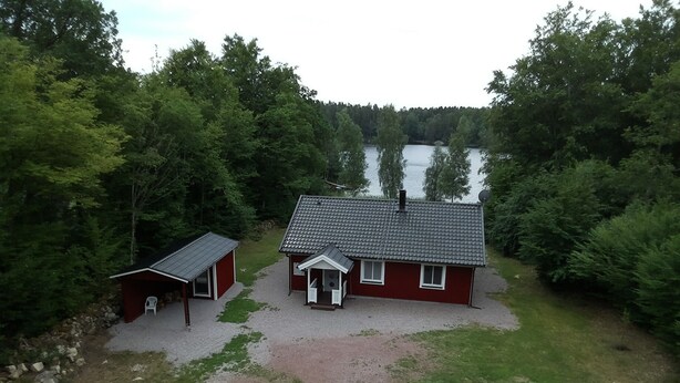Eine rote Holzhütte mit einem Ziegeldach, einer kleinen Veranda und einer Schotterauffahrt, die zu einem hölzernen Steg am See führt.