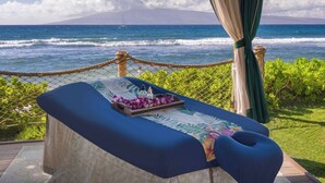 Ein Massagetisch mit floraler Abdeckung, eine Flasche und eine Schale mit Blumen, mit Blick auf den Ozean, inklusive Seilabsperrung und Grünpflanzen.