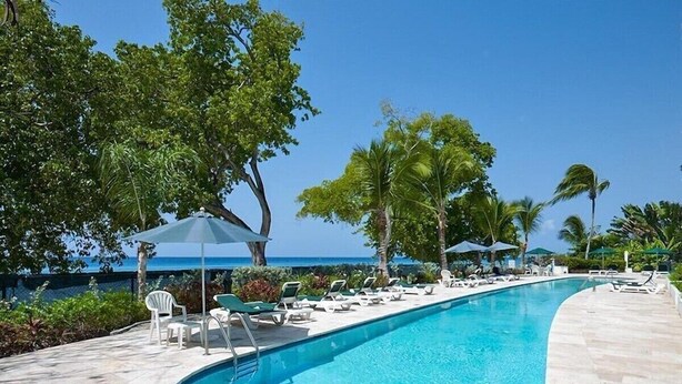 Une piscine entourée de chaises longues et de parasols, bordée de palmiers et surplombée d’un ciel dégagé.