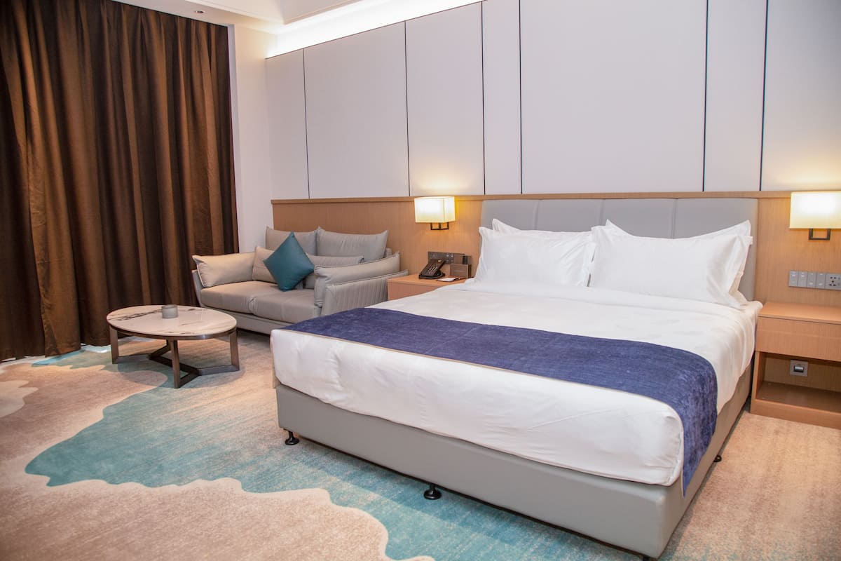 A hotel room with a large bed, a sofa, a small round table, and a nightstand with a lamp.