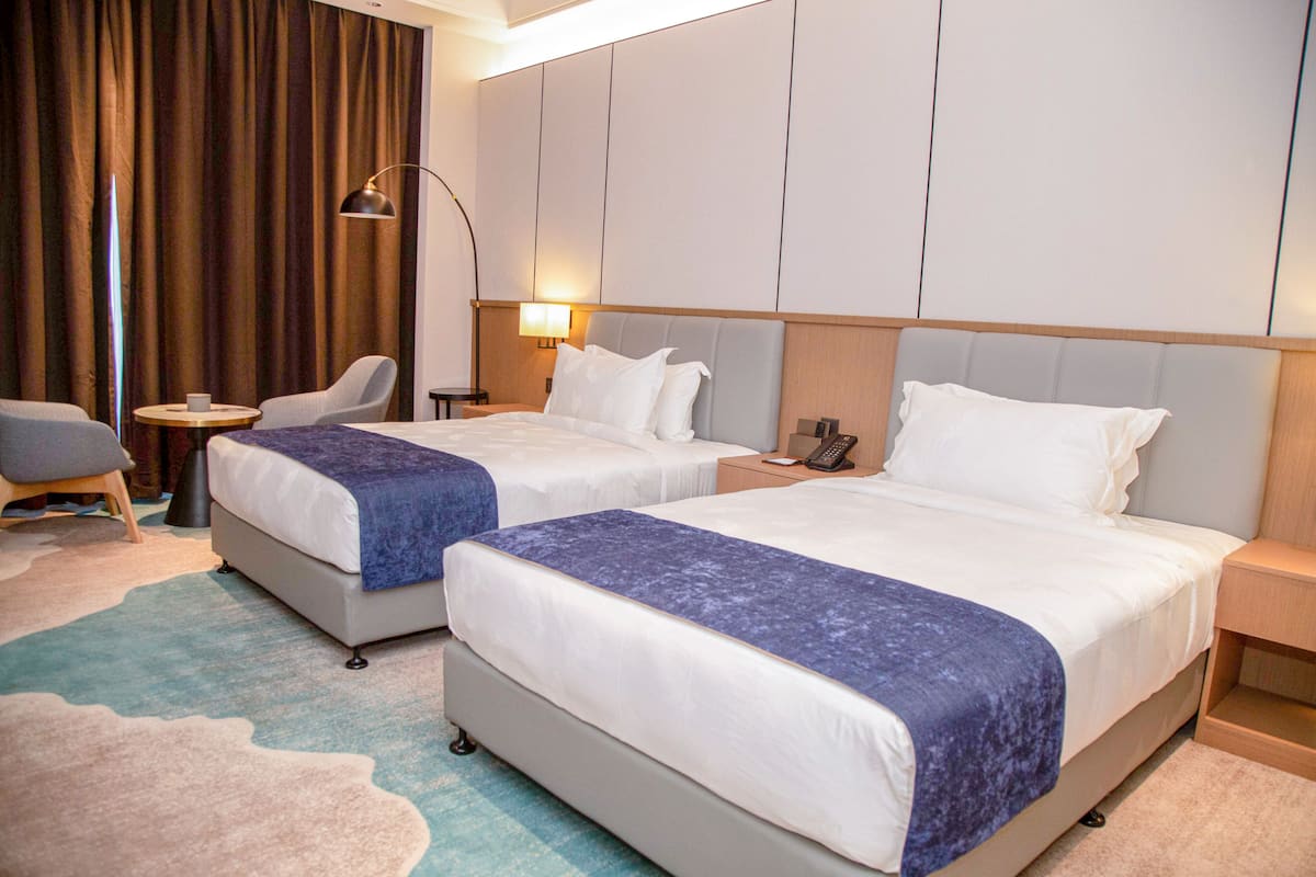 A hotel room with two beds, a desk, a chair, a lamp, and a large window with curtains.