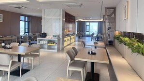 A modern dining area with multiple tables and chairs, a kitchen area with a buffet, and a view of the city through large windows. - LC International Hotel (Yangon)