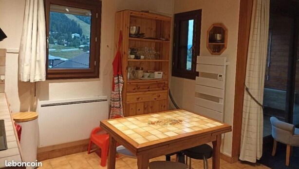 Ein gemütliches Zimmer mit einem Holztisch, einem roten Hocker, einer Speisekammer mit Geschirr und einem Fenster mit Bergblick.